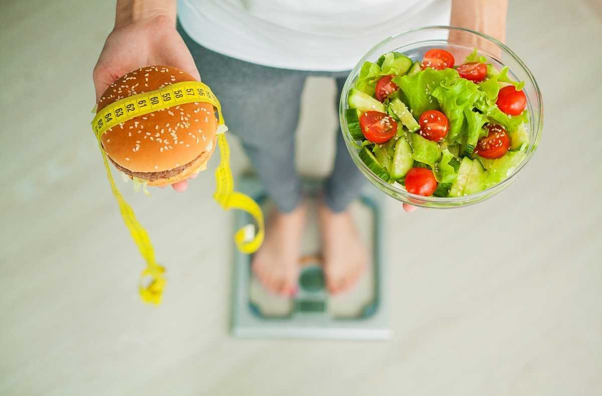 Pessoa sobre a balança segurando um hambúrguer na mão direita e um prato de salada na mão esquerda