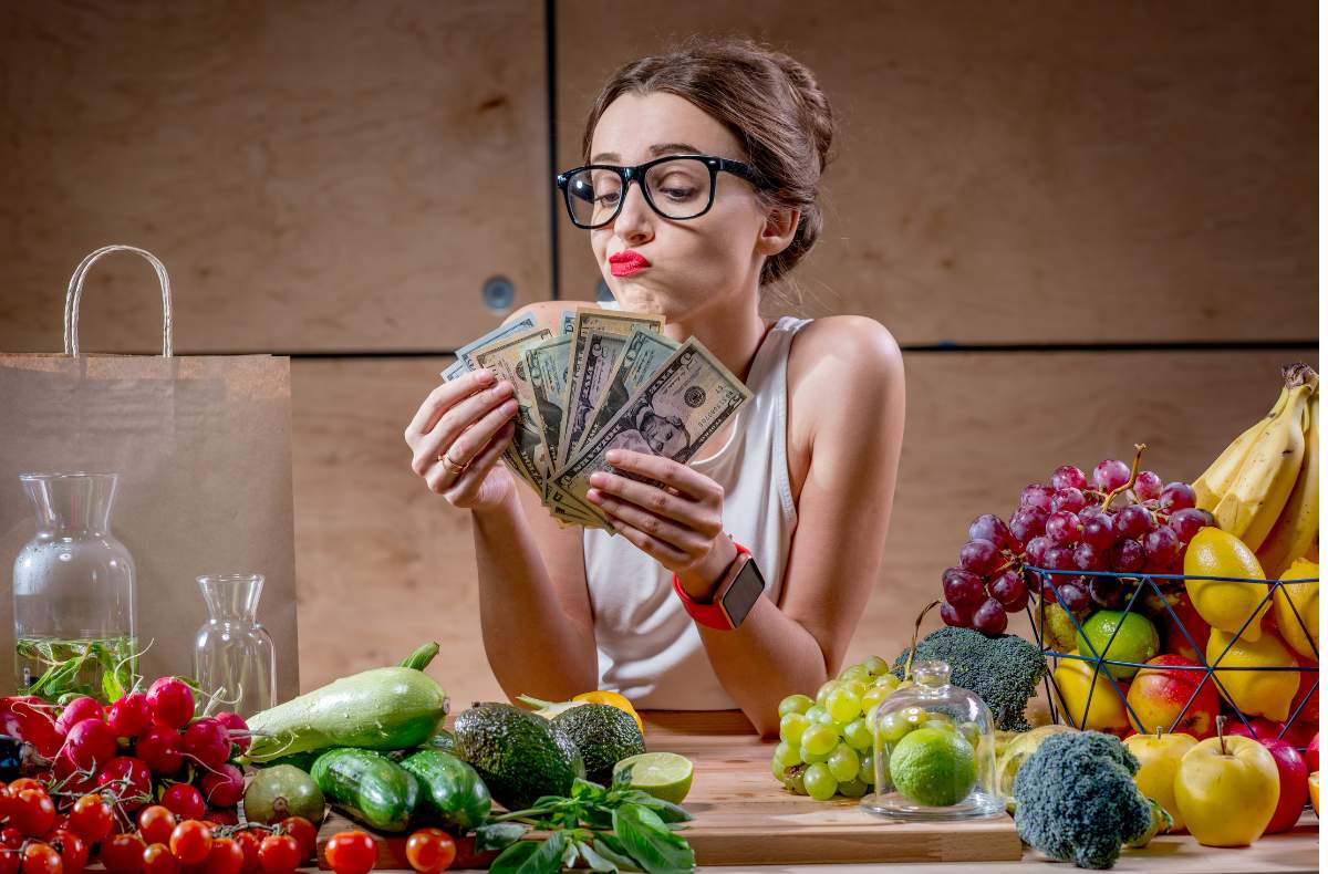 Mulher contando dinheiro, sentada com os cotovelos apoiados sobre uma mesa que está cheia de alimentos da feira.